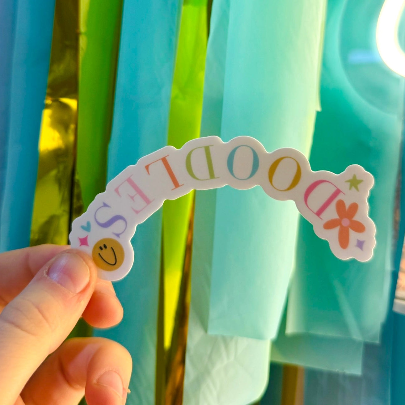 Doodles by Rebekah arched "DOODLES" logo vinyl sticker. This whimsical hand-painted brand decal features pastel typography, flowers, and a smiley face, making it a perfect decorative accent for water bottles, laptops, and craft room planners.