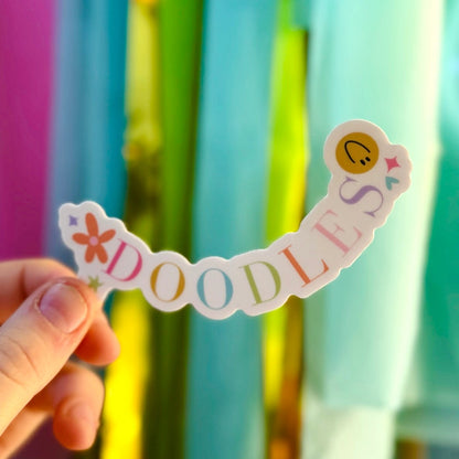A hand holds a die-cut vinyl sticker curved into a smile shape against a background of festive pink, teal, and lime green streamers. The sticker features the word "DOODLES" in soft pastel colors, with a hand-painted orange flower on the left and a yellow smiley face with pink and blue hearts on the right.
