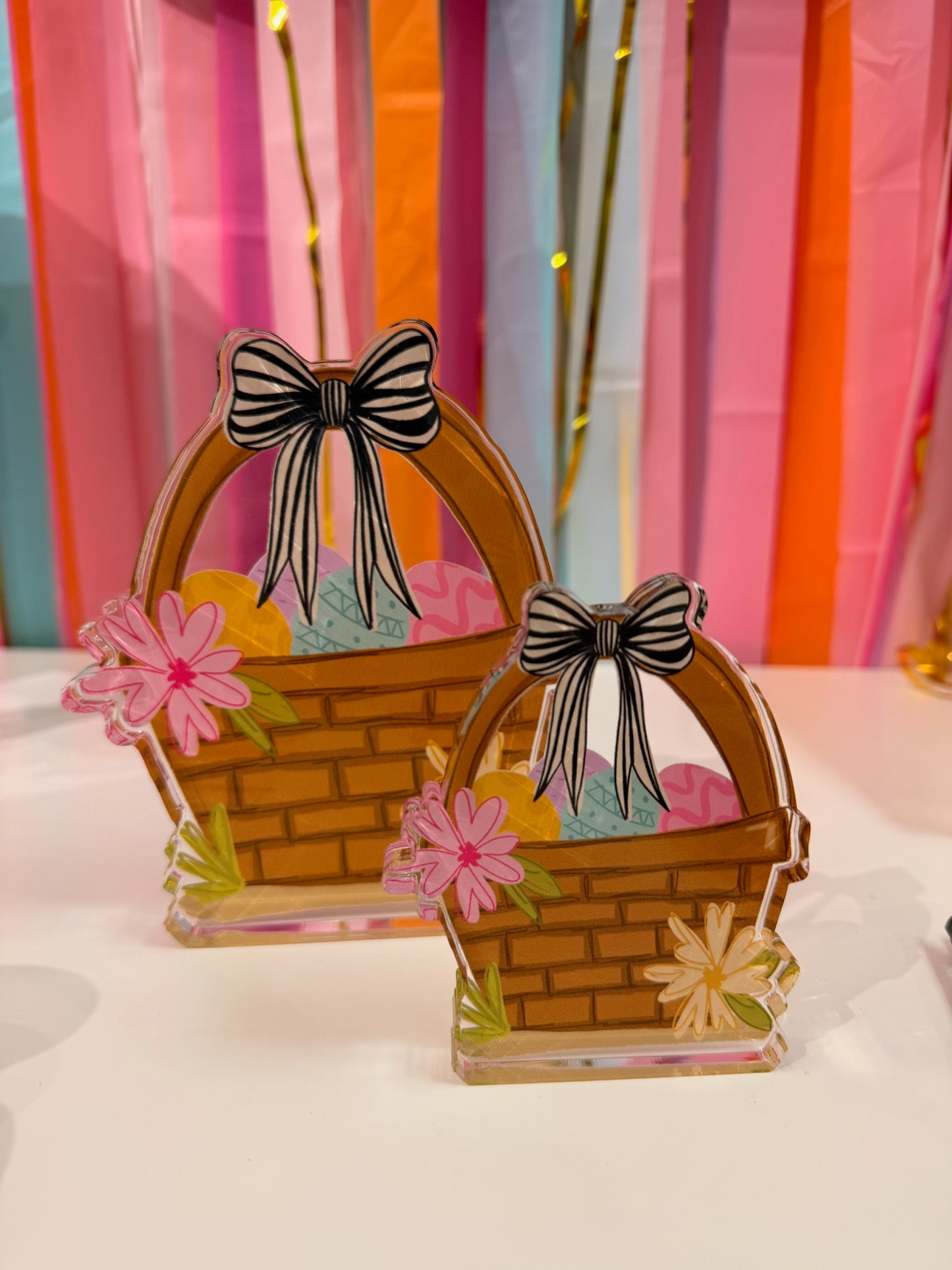 Doodles by Rebekah Easter basket acrylic home decor featuring whimsical hand-painted eggs, a striped bow, and floral details. These colorful spring tabletop accents are shown against a bright party streamer background.