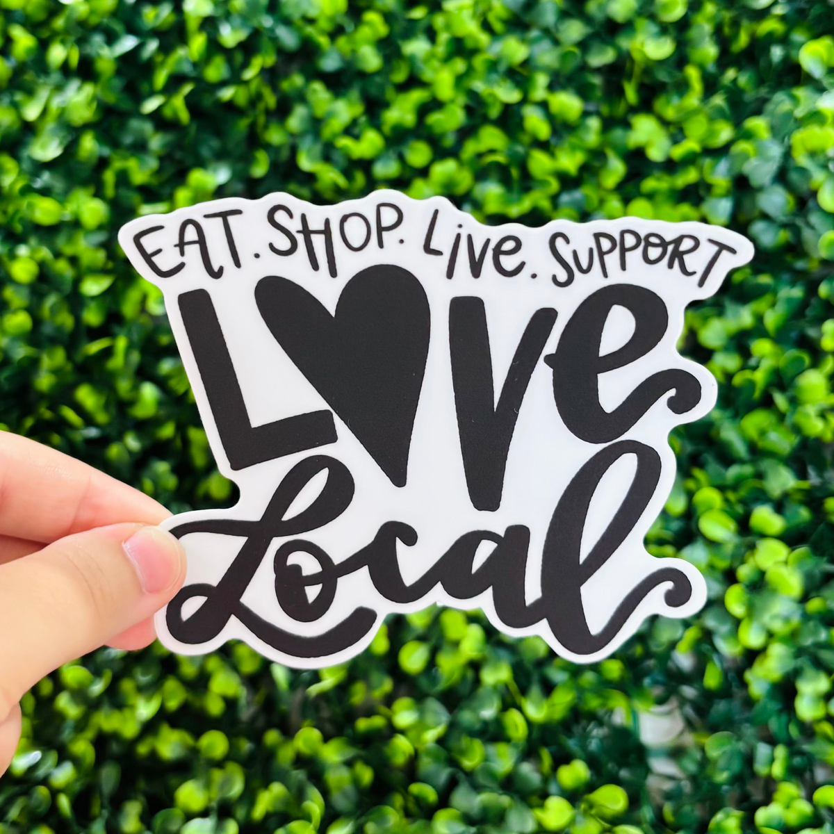A hand holding a black and white sticker that reads "Eat. Shop. Live. Support. Love Local"