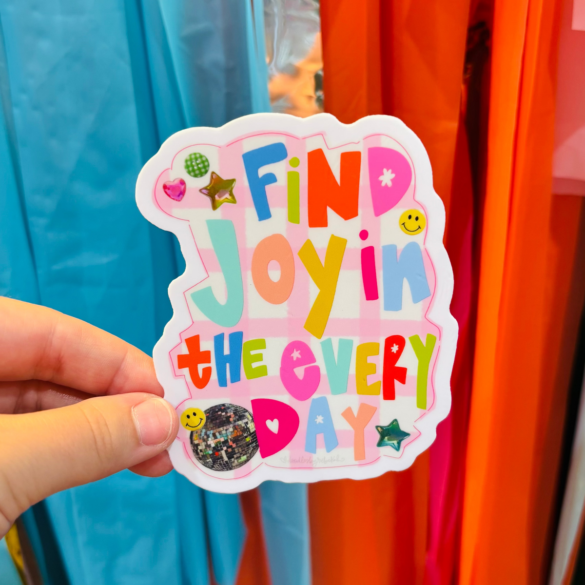 a hand holding a sticker that reads "Find Joy In the Everyday" with a decorative background