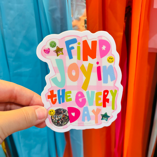 a hand holding a sticker that reads "Find Joy In the Everyday" with a decorative background