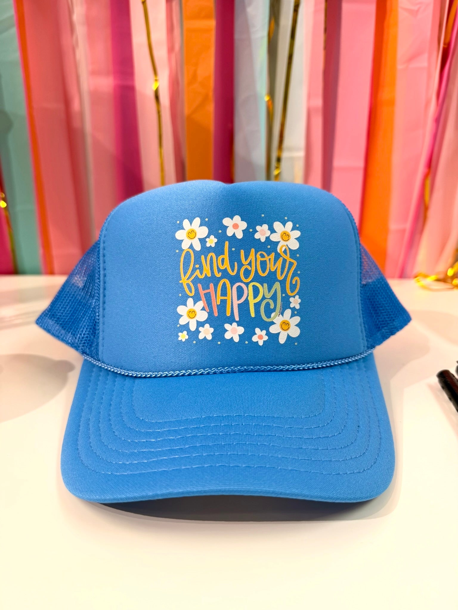 Blue "Find Your Happy" trucker hat by Doodles by Rebekah featuring whimsical hand-drawn typography and smiley face daisies on a colorful streamer background.