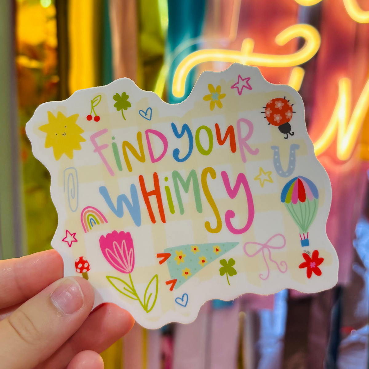 a hand holding a colorful hand draw sticker that reads "Finds Your Whimsy"