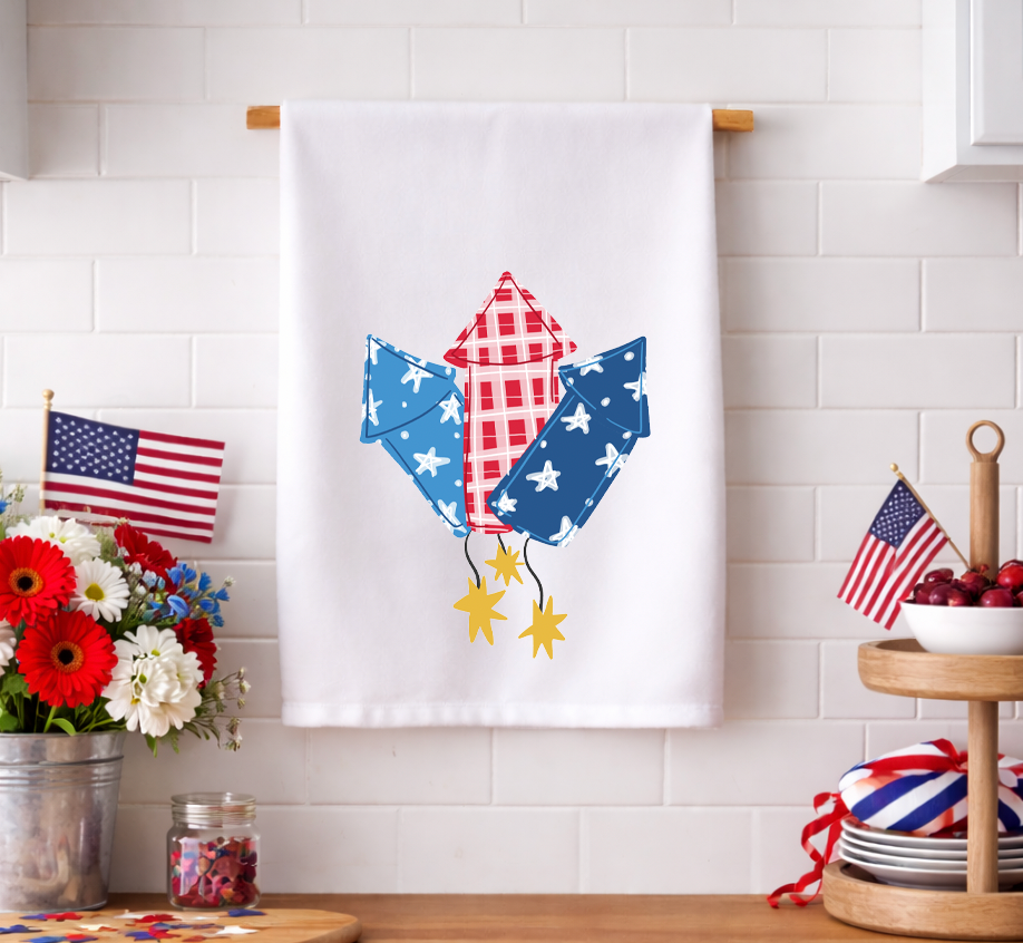 Patriotic 4th of July decorative kitchen tea towel featuring whimsical red, white, and blue firework rocket illustrations, displayed in a festive kitchen setting with American flags.