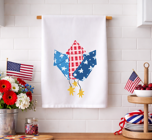 Patriotic 4th of July decorative kitchen tea towel featuring whimsical red, white, and blue firework rocket illustrations, displayed in a festive kitchen setting with American flags.