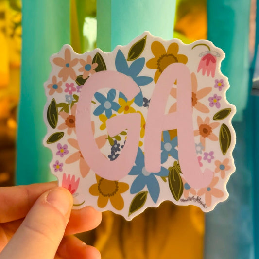 Doodles by Rebekah Georgia "GA" state pride floral sticker. This whimsical hand-painted vinyl decal with botanical accents and pink lettering is a perfect gift for Georgians to decorate water bottles, laptops, and planners.