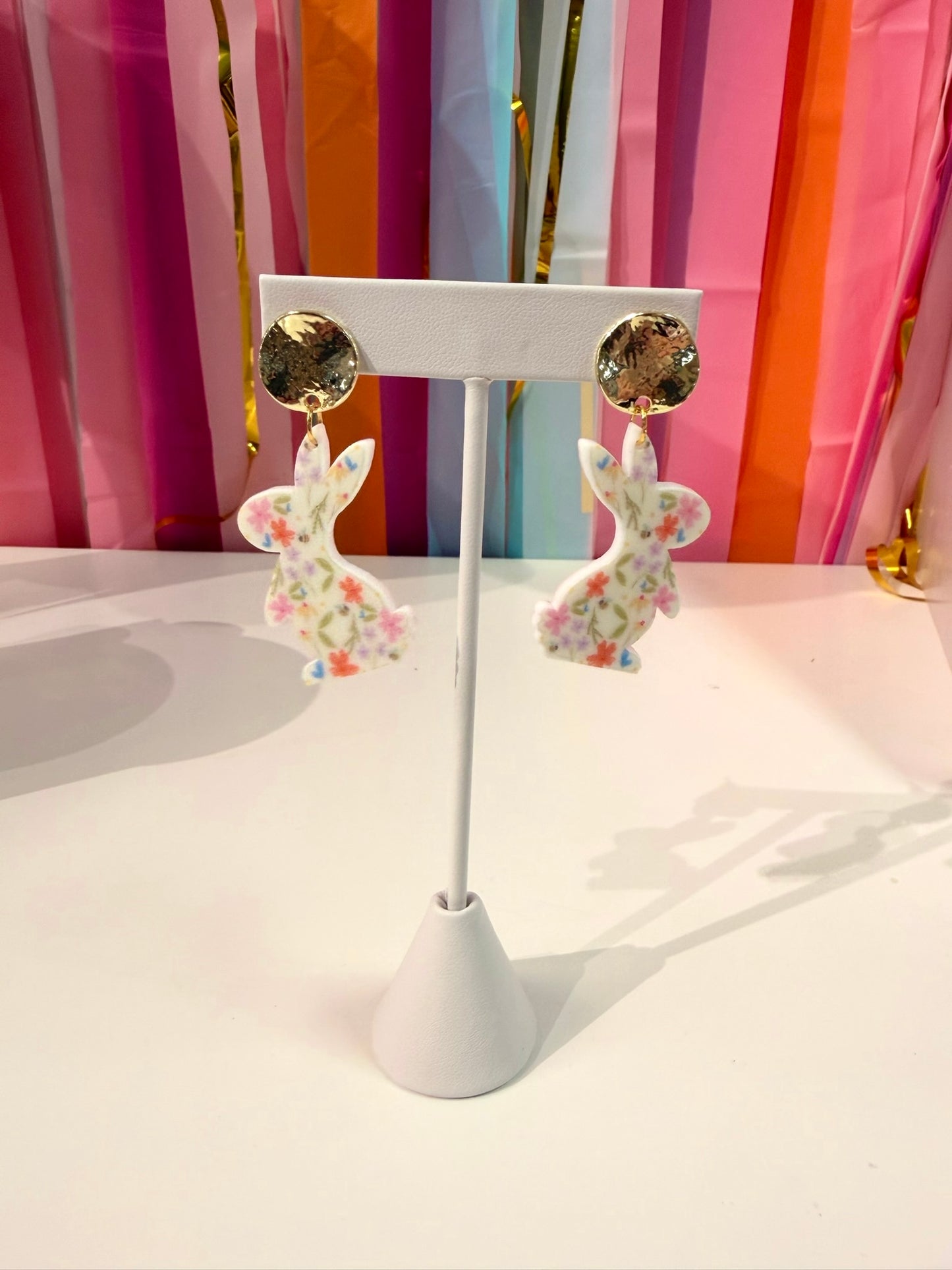 Doodles by Rebekah floral bunny statement earrings featuring hand-drawn spring wildflower patterns and hammered gold studs. These colorful Easter-themed accessories are shown against a bright party streamer backdrop.