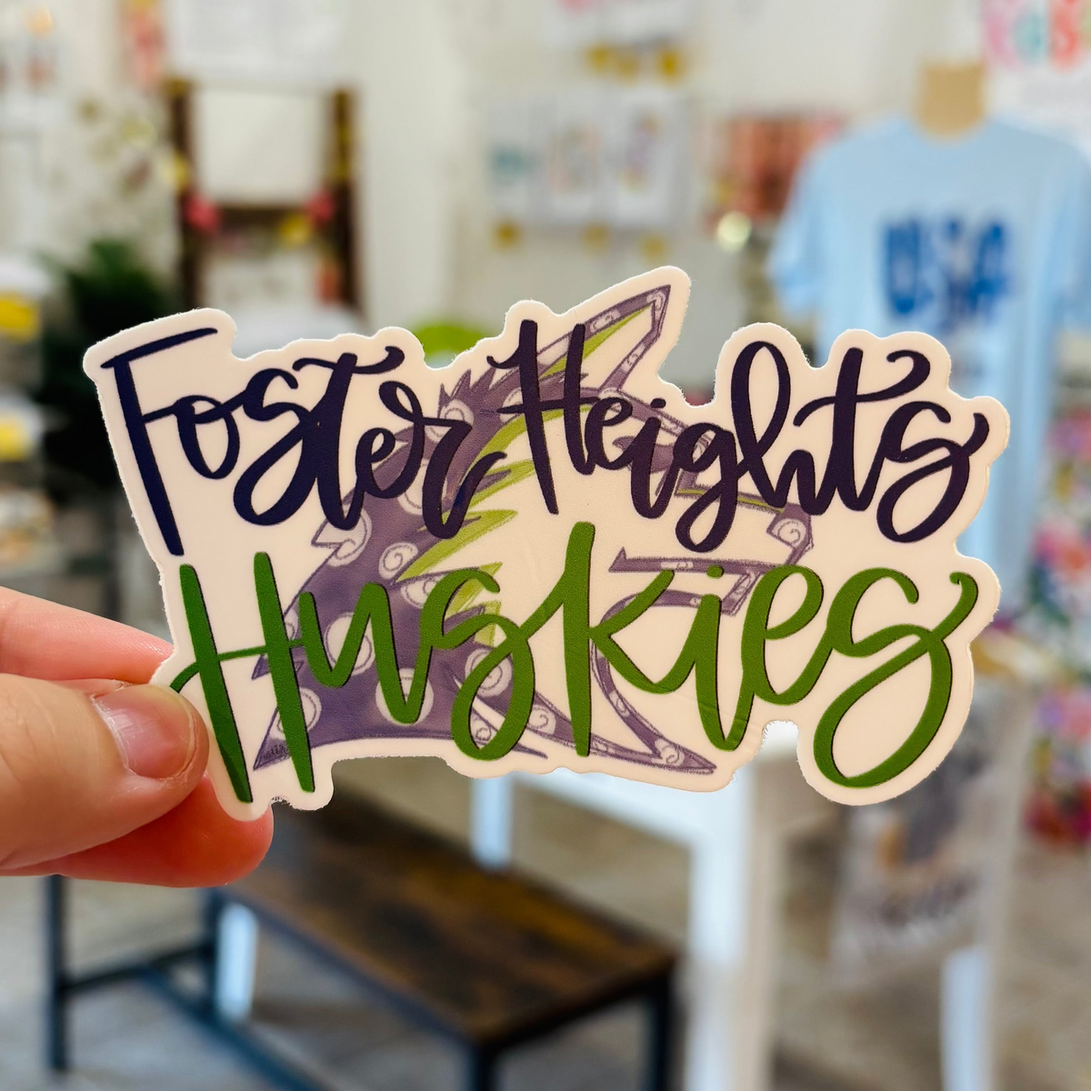 a hand holding a sticker that reads "Foster Heights Huskies" with a purple & green husky in the background
