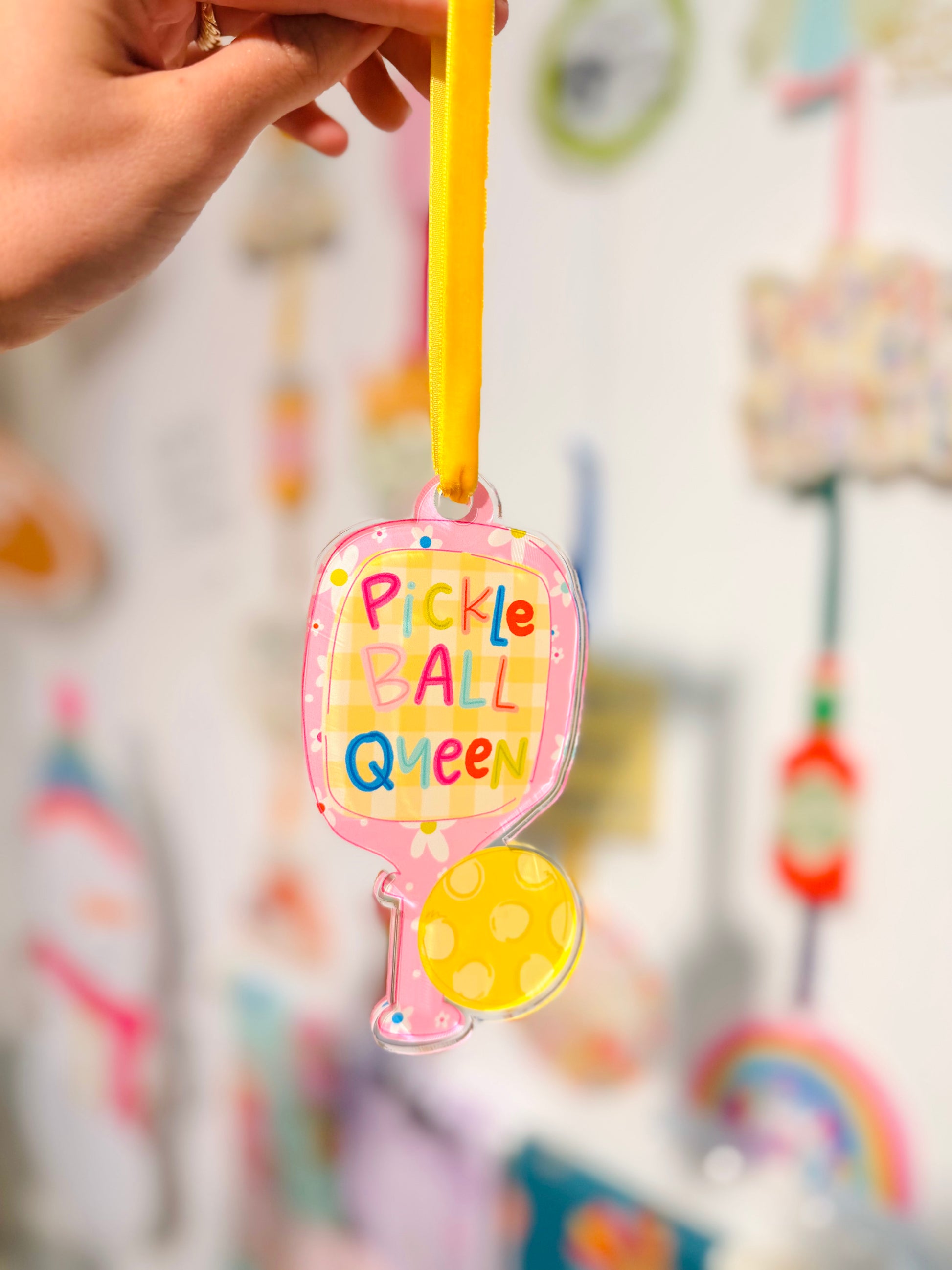 Colorful 'Pickle Ball Queen' ornament held by a hand with a blurred background of colorful items.