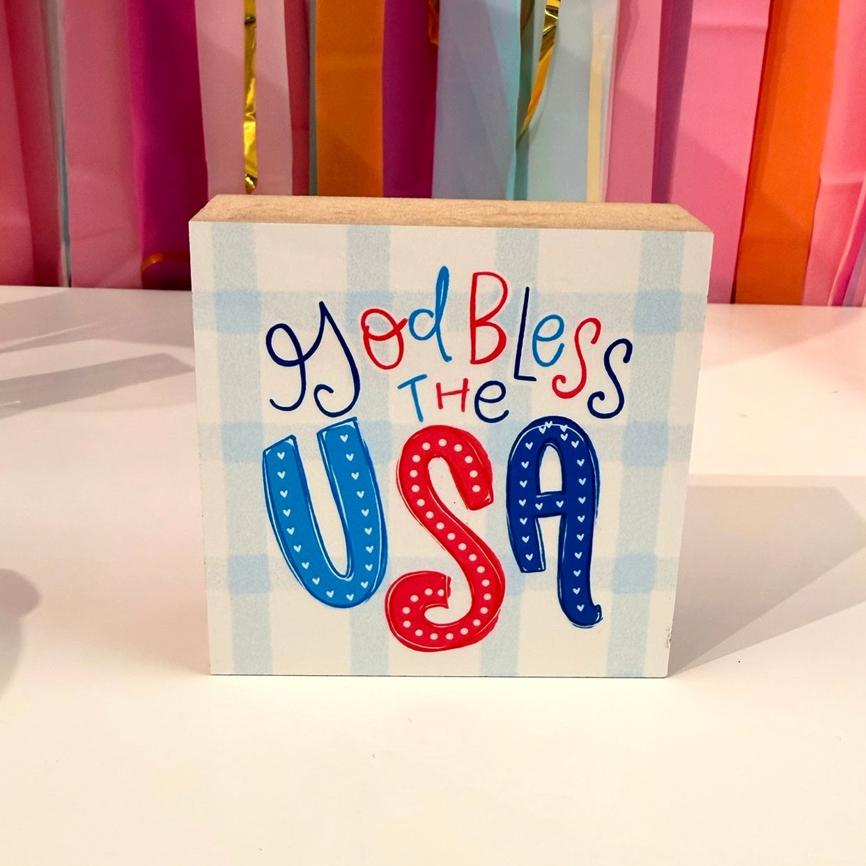 Doodles by Rebekah patriotic wooden block home decor featuring "God Bless the USA" hand-painted typography on a light blue checkered pattern. This whimsical red, white, and blue tabletop or shelf accent is shown against a bright party streamer background.