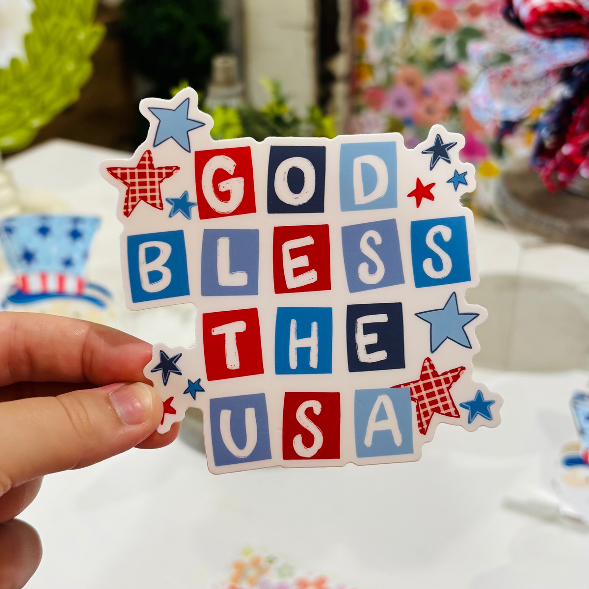 a hand holding a red, white, and blue sticker with blocky letters that reads "God Bless The USA"
