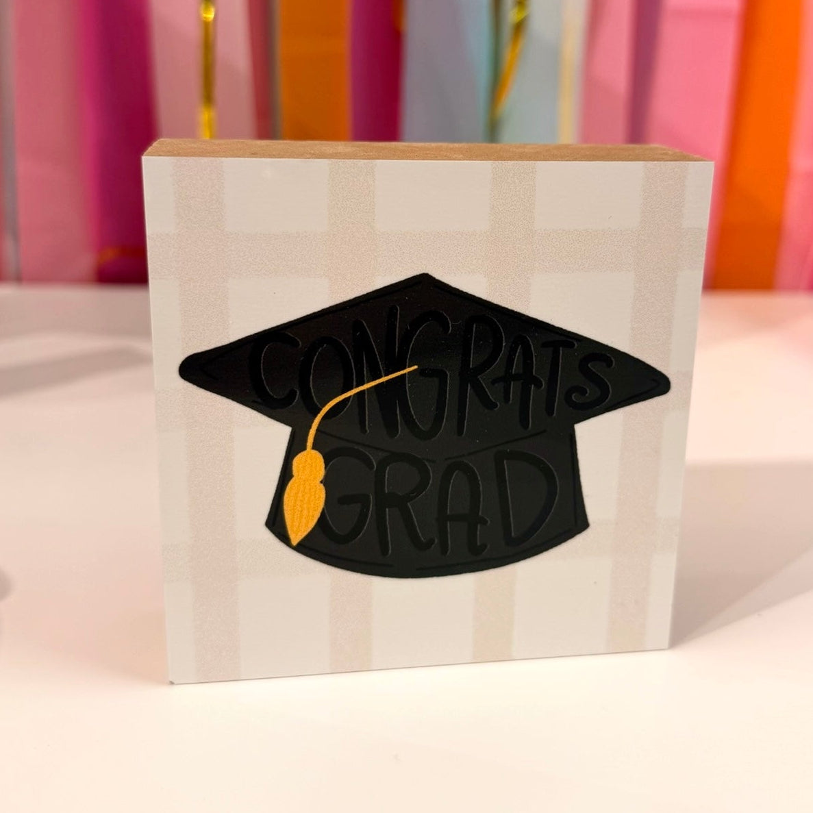 Doodles by Rebekah graduation wooden block home decor featuring a hand-painted black "Congrats Grad" mortarboard with a gold tassel. This whimsical commemorative gift or tabletop graduation party accent is shown against a bright streamer background.
