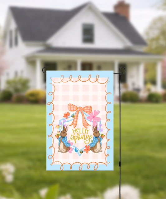 Doodles by Rebekah "Hello Spring" garden flag featuring whimsical hand-painted rabbits and an orange checkered bow. This charming Easter and spring-themed outdoor decor, with its peach checkered pattern and blue border, is perfect for front yards or garden displays.