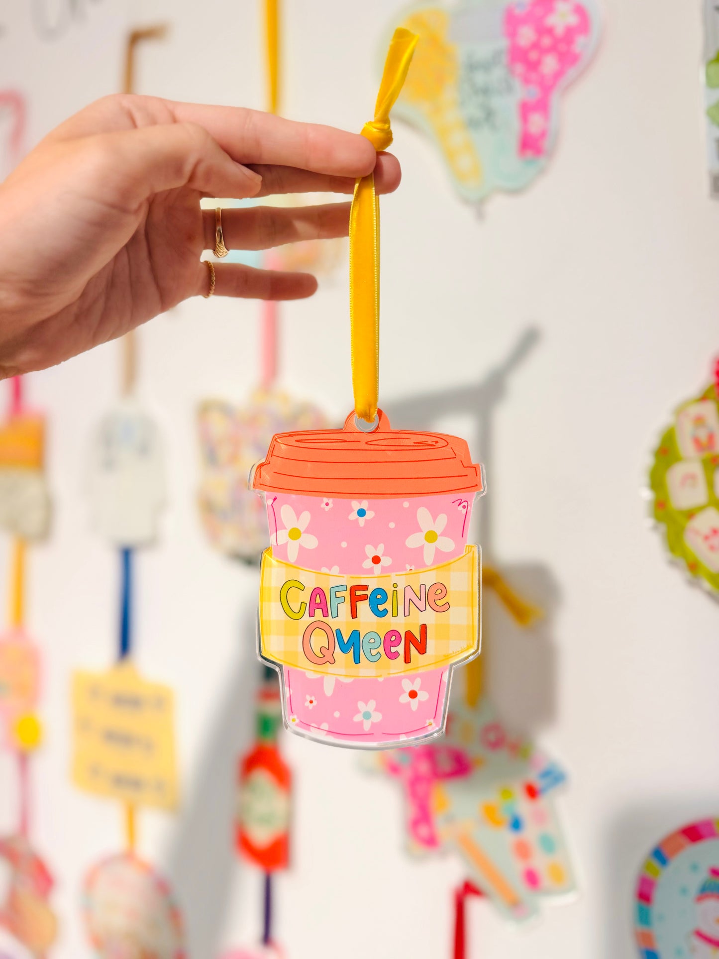 Pink drink cup ornament with 'Caffeine Queen' text held by a hand against a colorful background.