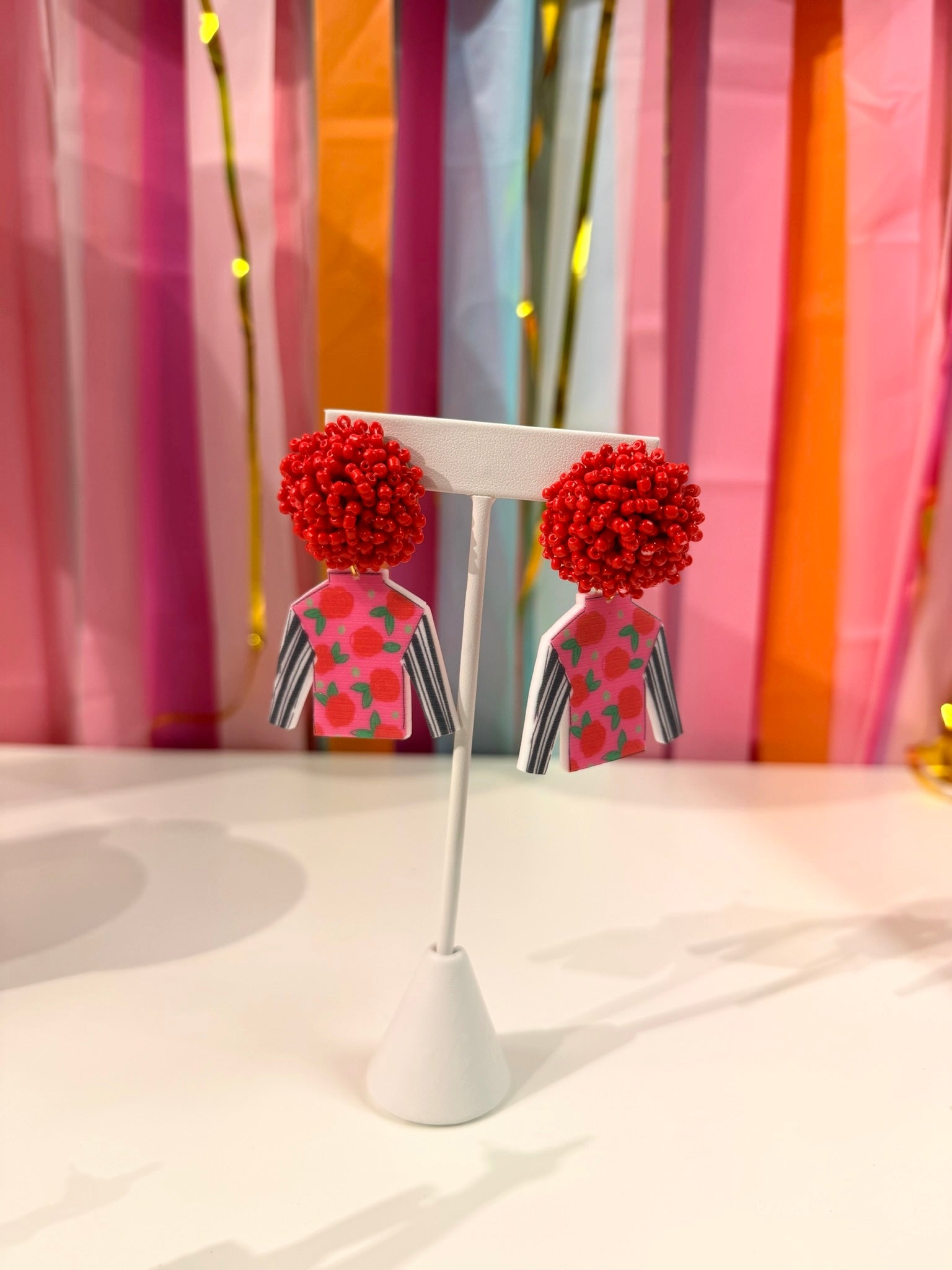 Doodles by Rebekah Kentucky Derby jockey silk statement earrings featuring hand-drawn red rose patterns, striped sleeves, and red beaded studs. These whimsical horse racing accessories are shown against a bright party streamer backdrop.