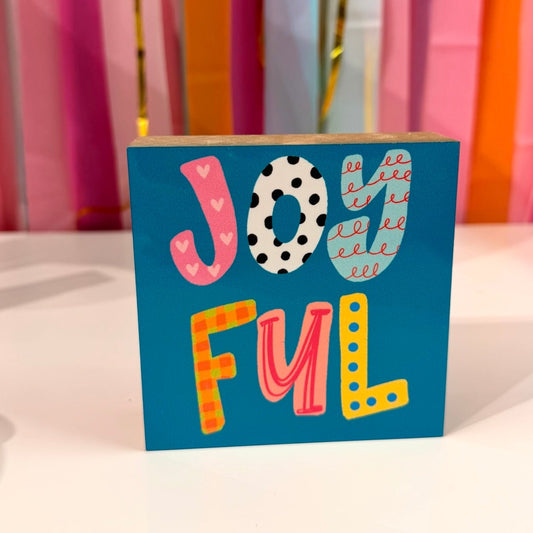 Doodles by Rebekah "Joyful" wooden block home decor featuring whimsical, hand-painted patterned typography on a teal background. This bright and cheerful tabletop or shelf accent is shown against a colorful party streamer backdrop.
