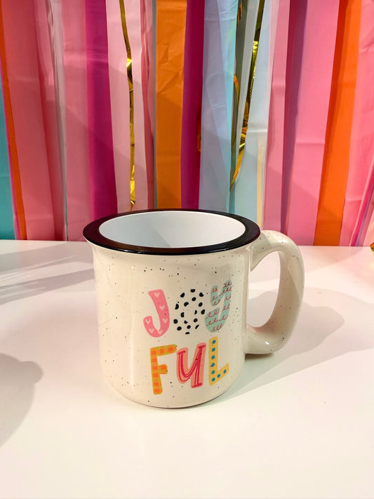 Doodles by Rebekah "JOYFUL" hand-painted campfire mug with colorful patterned typography. This whimsical ceramic coffee cup is displayed against a festive streamer backdrop, making it an ideal gift for birthdays, celebrations, or as a cheerful addition to kitchen decor.