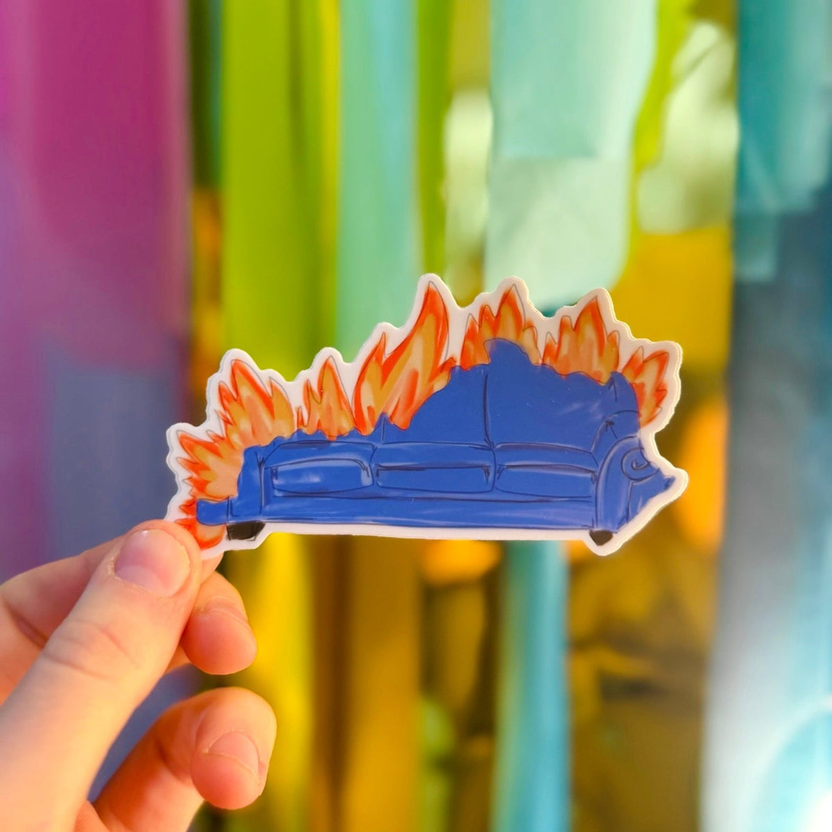 Doodles by Rebekah burning couch sticker. This whimsical vinyl decal features a hand-painted blue sofa on fire, making it a humorous gift or a unique accessory for laptops and water bottles, often associated with college sports traditions or celebrations.