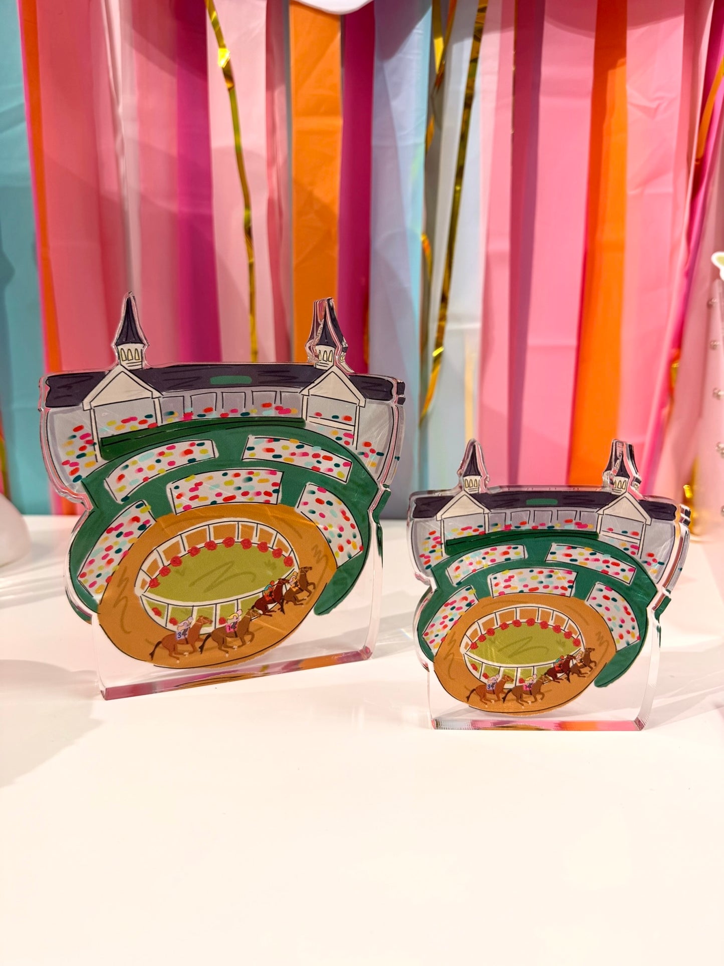Doodles by Rebekah Kentucky Derby racetrack acrylic home decor featuring whimsical hand-painted grandstands and the iconic twin spires. These colorful horse racing tabletop accents are perfect for Derby Day celebrations and are shown against a bright party streamer background.