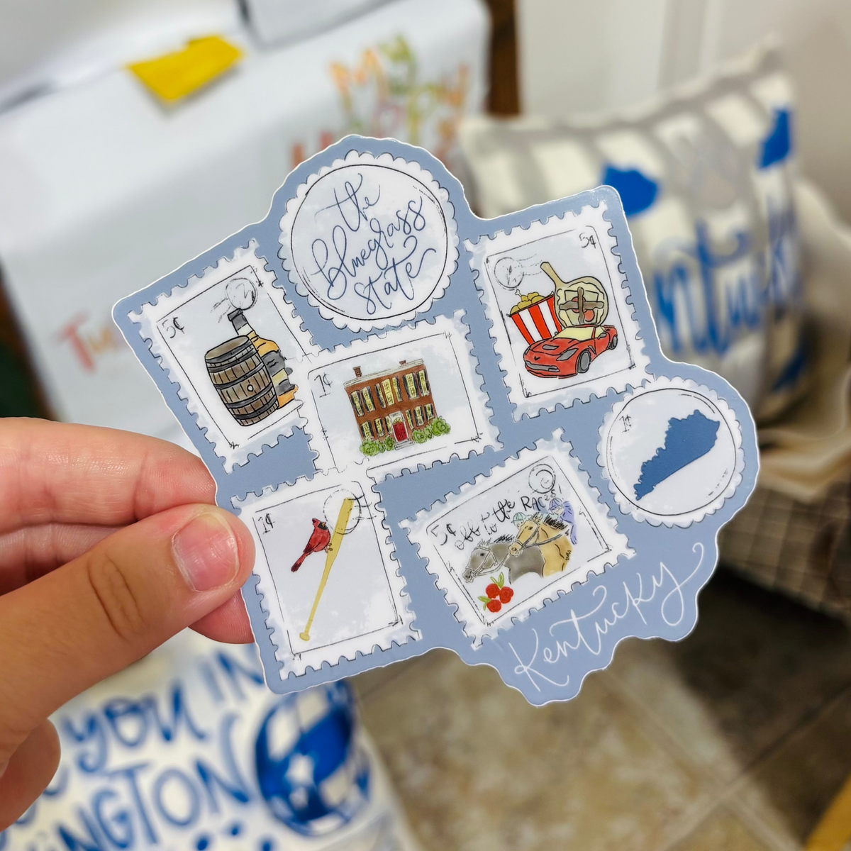 A hand holding a sticker of stamps representing the state of Kentucky on a blue background