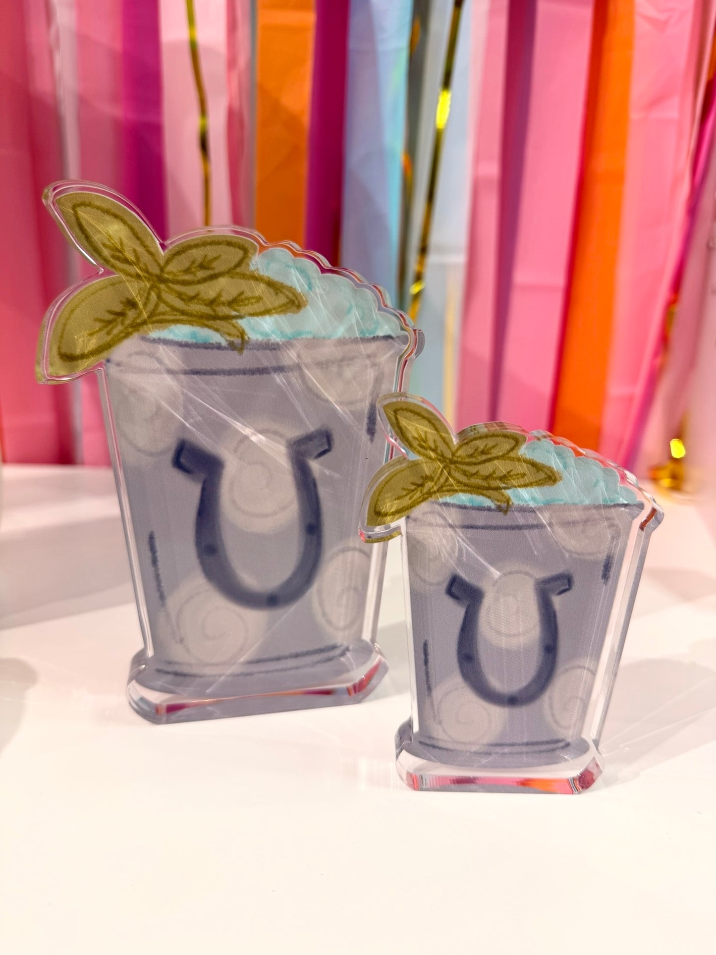 Doodles by Rebekah Kentucky Derby mint julep acrylic decor featuring a whimsical hand-painted cup design with horseshoe and mint leaf accents. These horse racing party decorations are shown against a colorful party streamer background.