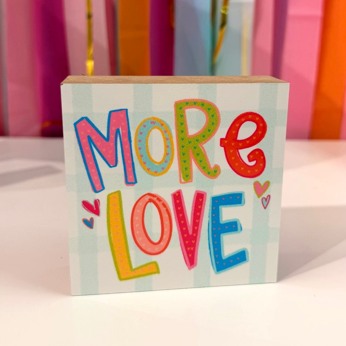 Doodles by Rebekah "More Love" wooden block home decor featuring multi-colored hand-painted patterned typography on a light blue checkered pattern. This cheerful and inspirational tabletop or shelf accent is shown against a bright party streamer background.