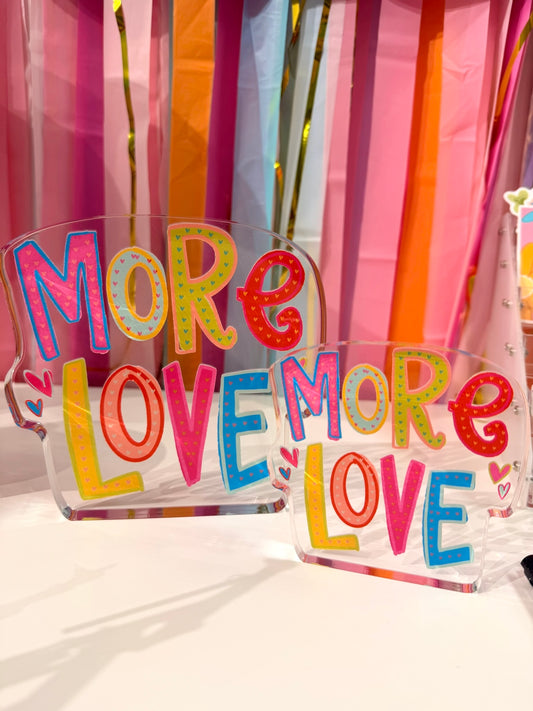 Doodles by Rebekah "More Love" acrylic home decor featuring whimsical hand-painted multi-colored typography and heart accents. These colorful Valentine's Day or everyday tabletop decorations are shown against a bright party streamer background.