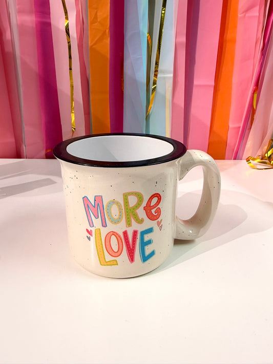 Doodles by Rebekah "More Love" hand-painted campfire mug featuring colorful patterned typography. This whimsical ceramic coffee cup is displayed against a festive streamer background, making it an ideal gift for Valentine’s Day, anniversaries, or positive home decor.