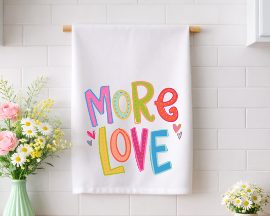 Whimsical "MORE LOVE" decorative tea towel with vibrant, hand-lettered typography and heart patterns, displayed in a bright kitchen with fresh floral arrangements.