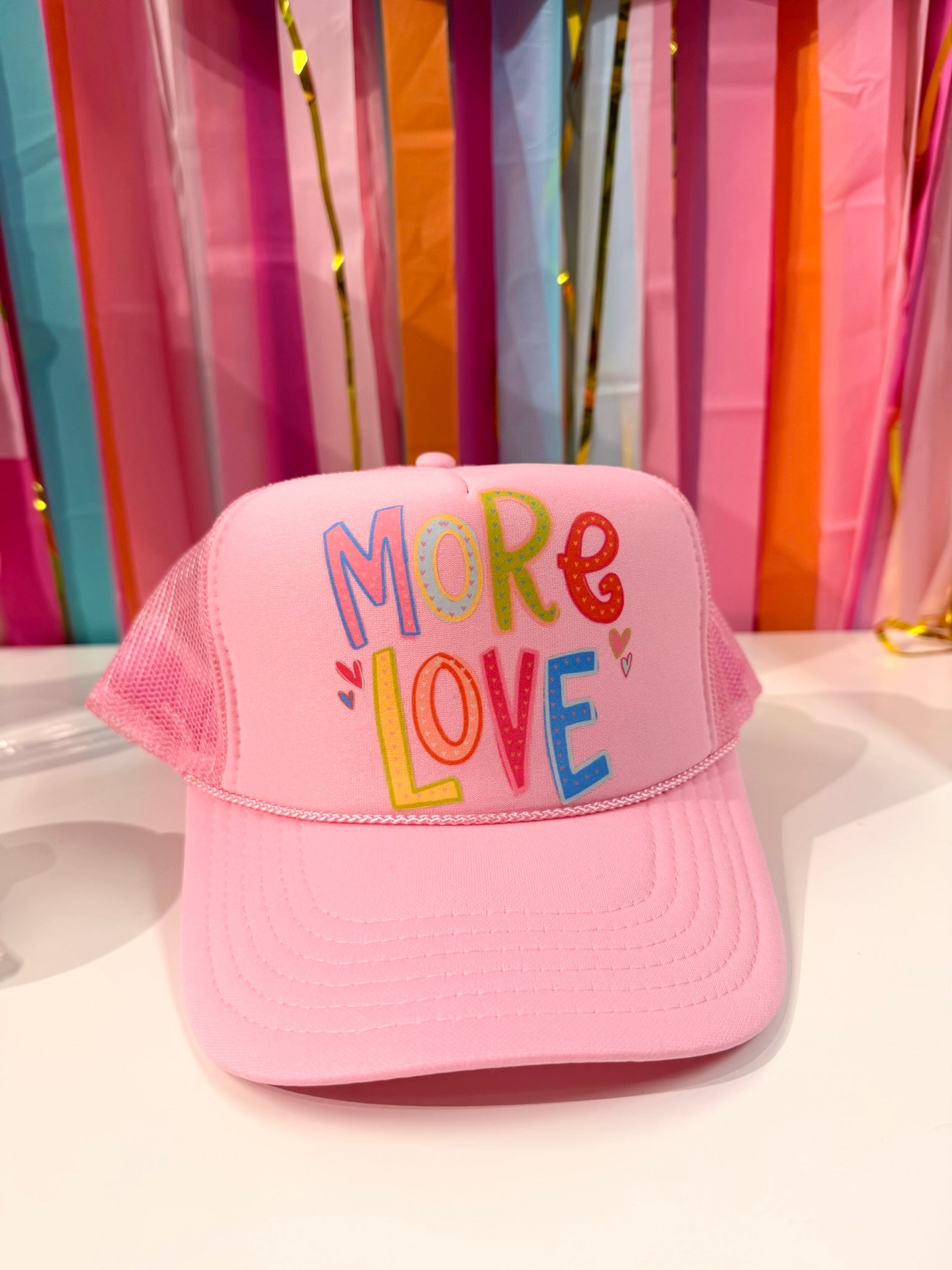 Pink "More Love" trucker hat from Doodles by Rebekah, featuring whimsical hand-drawn typography and heart illustrations on a colorful party streamer background.