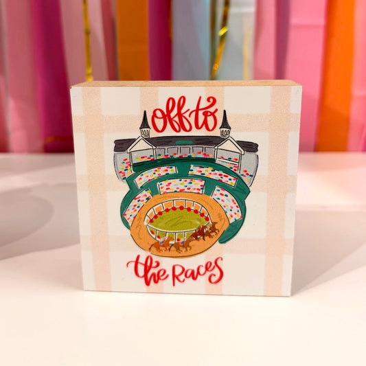 Doodles by Rebekah "Off to the Races" wooden block home decor featuring a whimsical hand-painted horse racing track and Churchill Downs-inspired spires. This equestrian-themed tabletop or shelf accent is shown against a bright party streamer background.