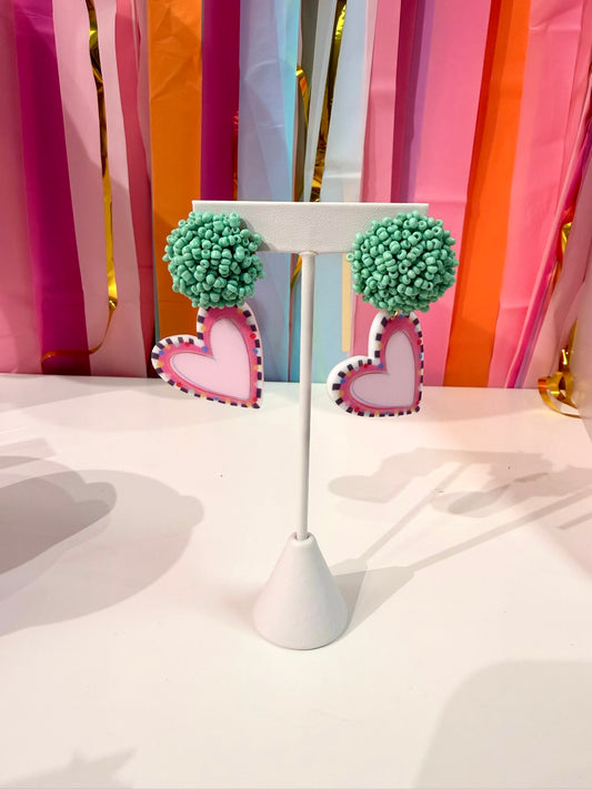 Doodles by Rebekah heart statement earrings featuring hand-painted pink hearts, checkered borders, and mint teal beaded studs. These whimsical Valentine’s Day or spring jewelry accessories are shown against a bright party streamer backdrop.