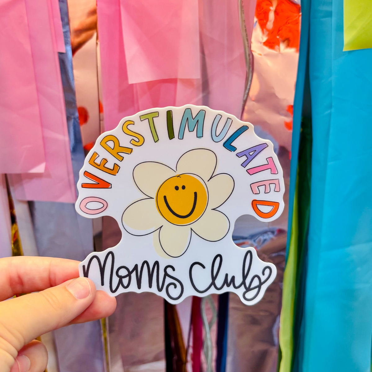 A hand holding a colorful sticker that reads Overstimulated Moms Club around a hand drawn daisy