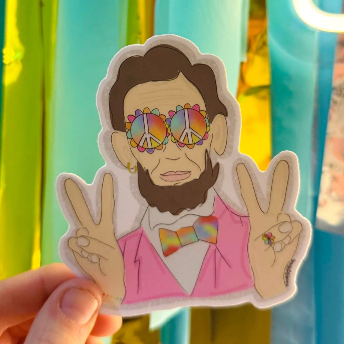 Doodles by Rebekah whimsical Abraham Lincoln sticker. This colorful, pop-art style historical figure decal features rainbow peace signs and a pink suit, making it a unique and fun gift for history buffs to use on water bottles, laptops, and journals.