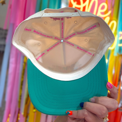 Teal cap with pink text held in front of colorful balloons and decorations