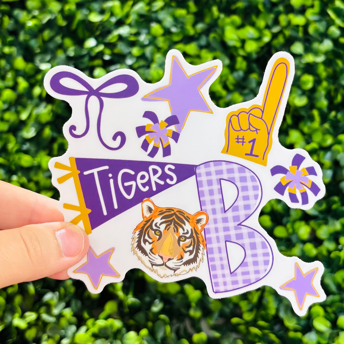 A hand holding a purple & yellow team Tigers sticker with game day inconography 