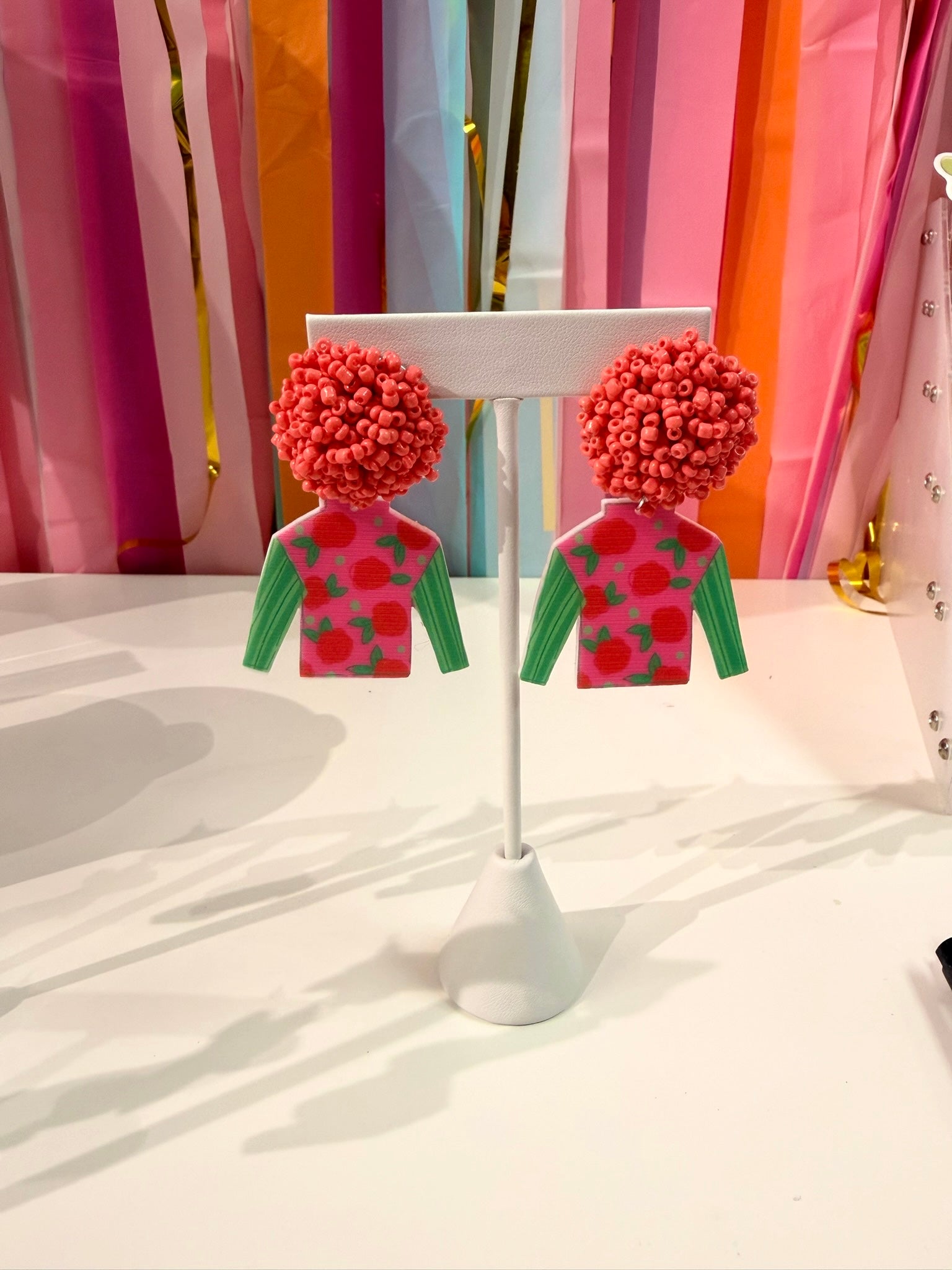 Doodles by Rebekah Kentucky Derby jockey silk earrings featuring hand-drawn rose patterns, green sleeves, and pink beaded studs. These whimsical horse racing accessories are shown against a bright party streamer backdrop.