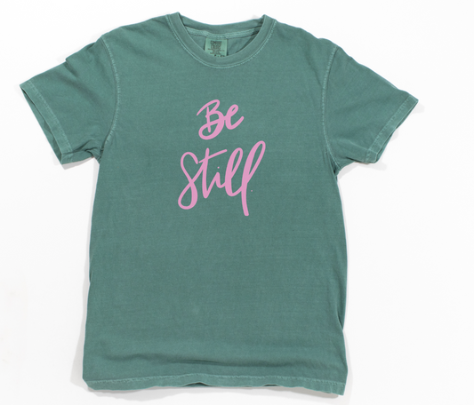 Green t-shirt with 'Be Still' text in pink on a white background