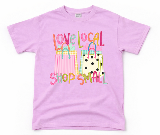 Pink t-shirt with colorful shopping bag graphics and 'love local shop small' text on a white background