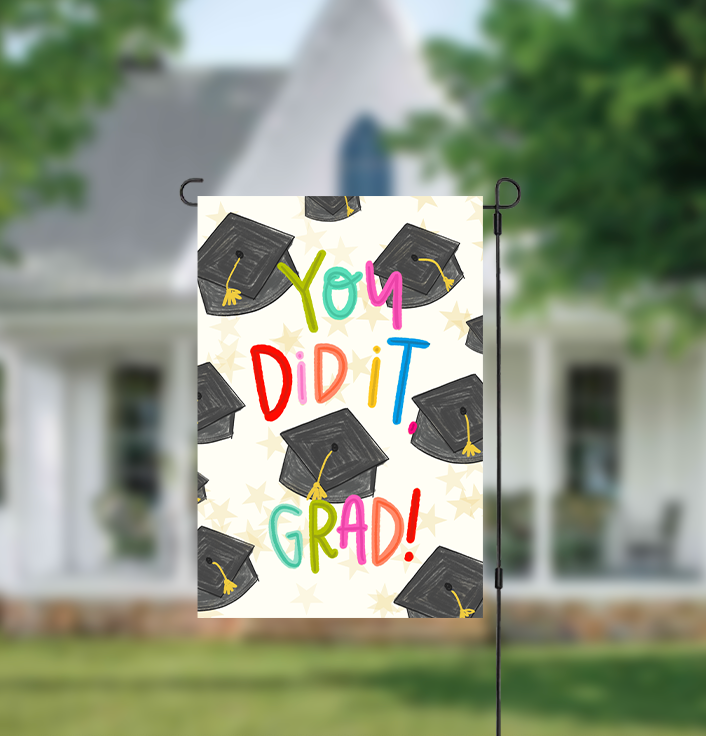 Graduation-themed garden flag with colorful text and caps on a blurred house background