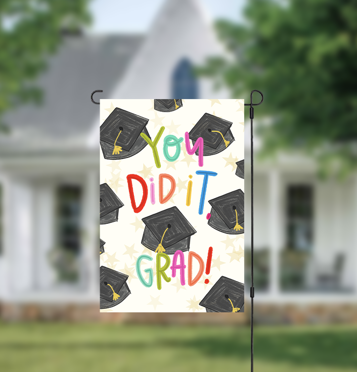 Graduation-themed garden flag with colorful text and caps on a blurred house background