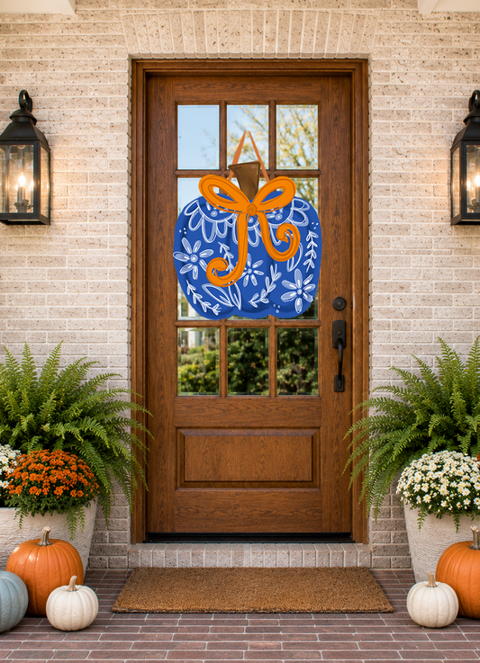 Decorative blue wreath with an orange bow on a wooden door with glass panels, flanked by potted plants and pumpkins.