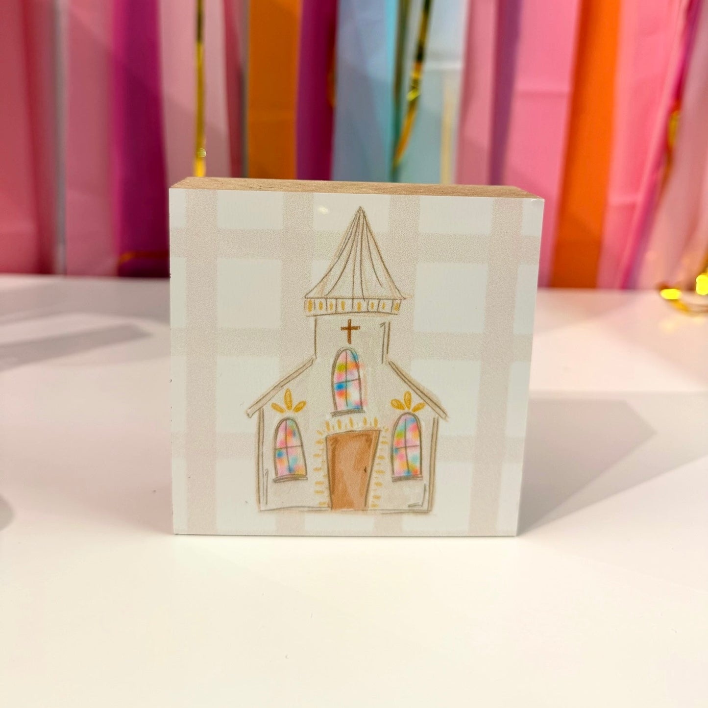 Doodles by Rebekah church wooden block home decor featuring a whimsical hand-painted country church on a neutral checkered pattern. This colorful tabletop or shelf accent is shown against a bright party streamer background.