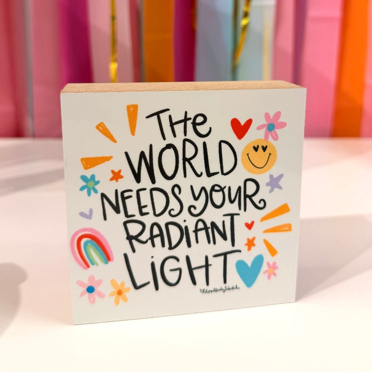 Doodles by Rebekah inspirational wooden block home decor featuring "The World Needs Your Radiant Light" whimsical hand-painted typography. This bright, positive tabletop or shelf accent is shown against a colorful party streamer background.