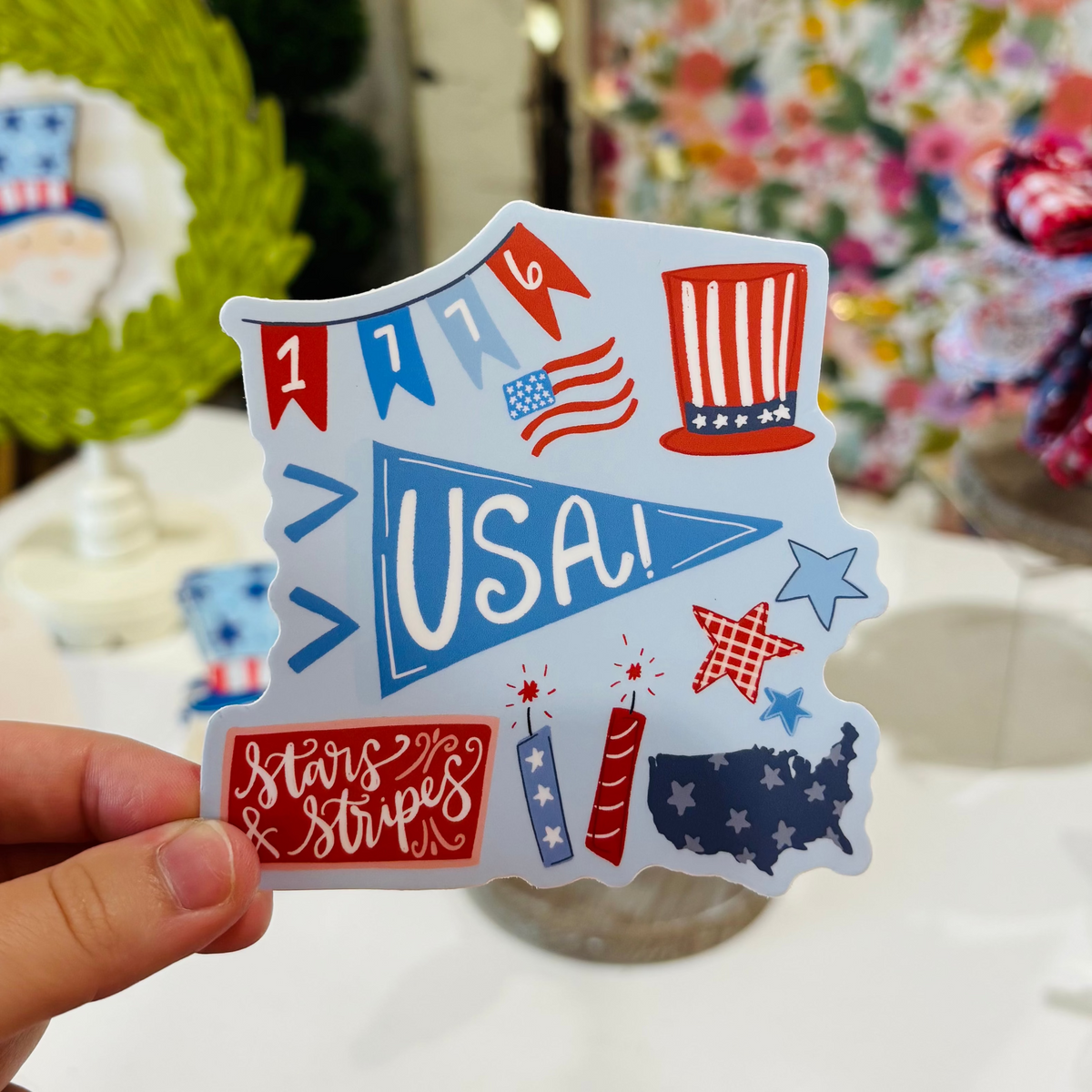 A Hand Holding a Sticker with a blue background and various Americana imagery in a hand drawn style 