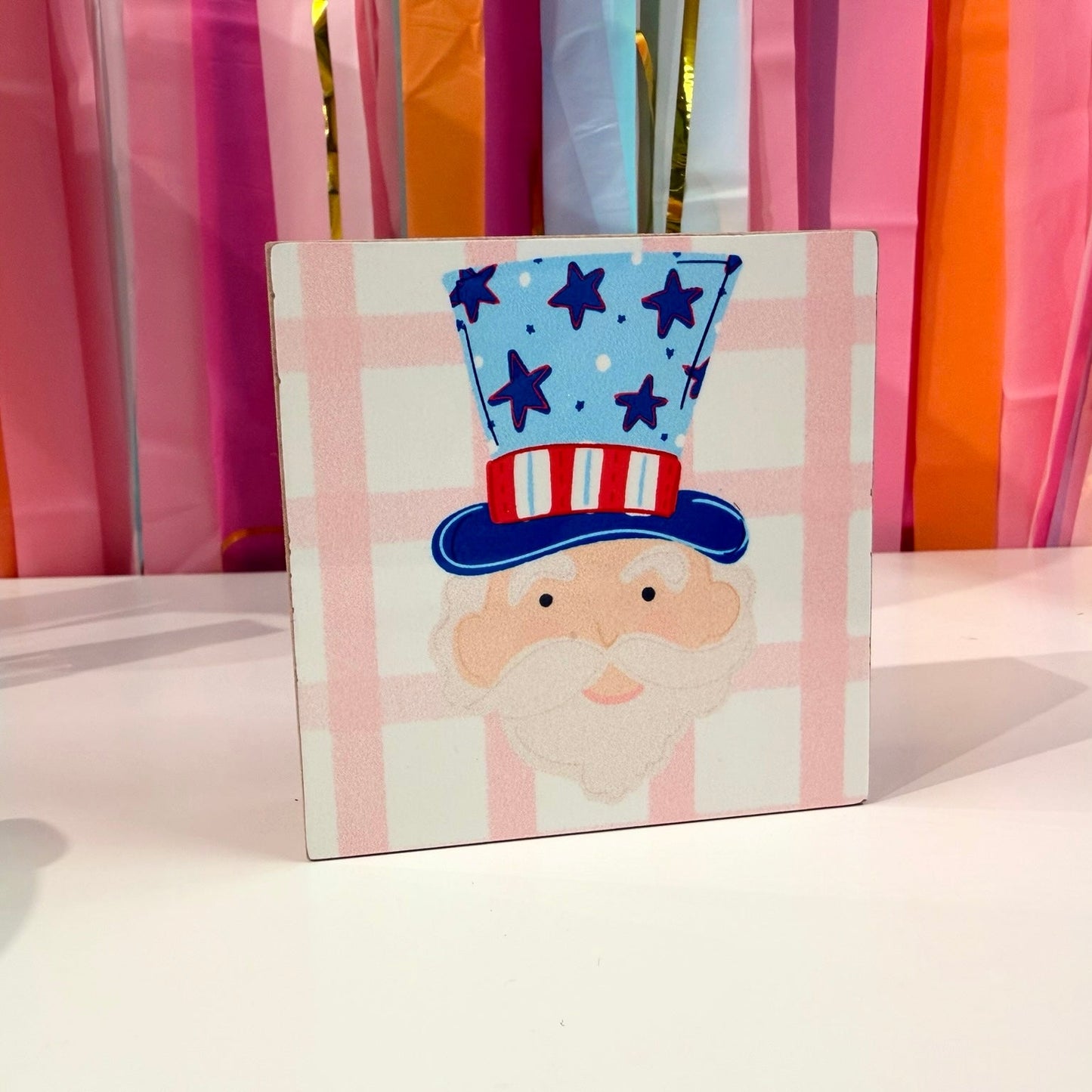 Doodles by Rebekah patriotic Uncle Sam wooden block home decor featuring a whimsical hand-painted design on a pink checkered pattern. This Americana-themed tabletop or shelf accent is shown against a bright party streamer background.
