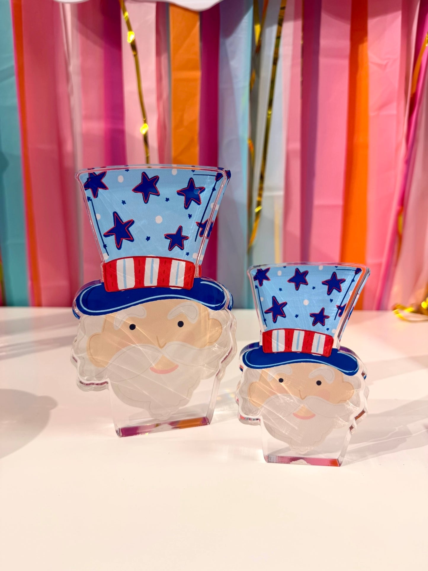 Doodles by Rebekah Uncle Sam acrylic home decor featuring a whimsical hand-painted patriotic design and starry top hat. These colorful Fourth of July tabletop accents are shown against a bright party streamer background.