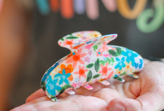 Hands cupping a tan hair claw clip featuring a whimsical orange, teal, and pink floral doodle pattern.