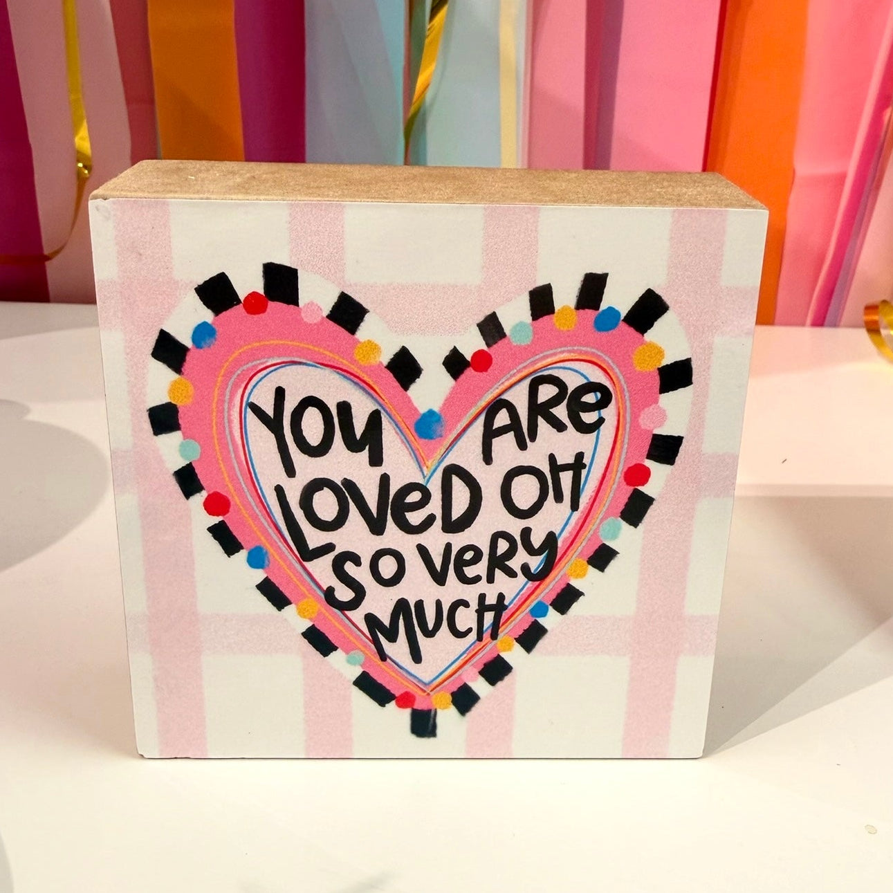 Doodles by Rebekah "You Are Loved" wooden block home decor featuring a whimsical hand-painted heart and checkered pattern. This colorful, inspirational tabletop or shelf accent is shown against a bright party streamer background.
